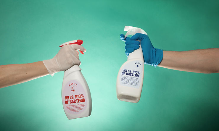 Two latex-gloved hands aiming spray bottles at one another with labels reading "kills 100% of bacteria."