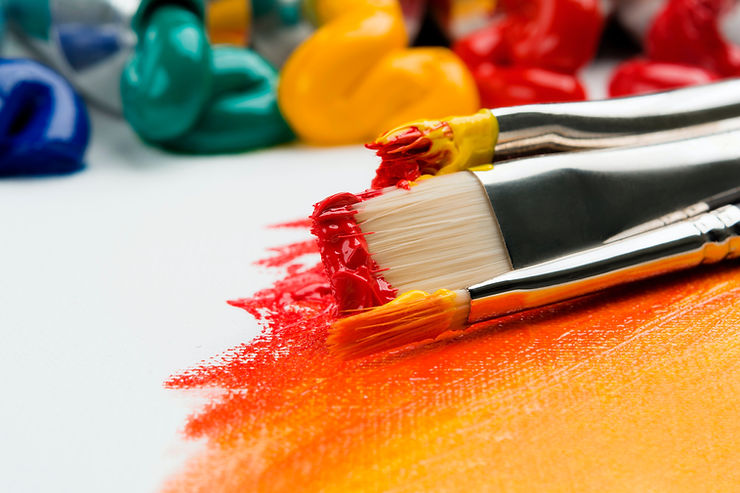 Two paint brushes loaded with red and yellow paint with tubes of paint squeezing out paint in the background.