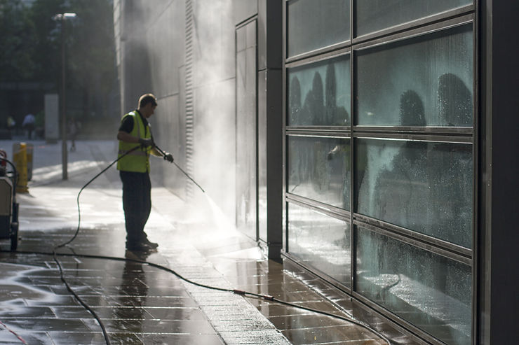 A man in a yellow vest spraying sidewalks in front of a glass and metal commercial building.