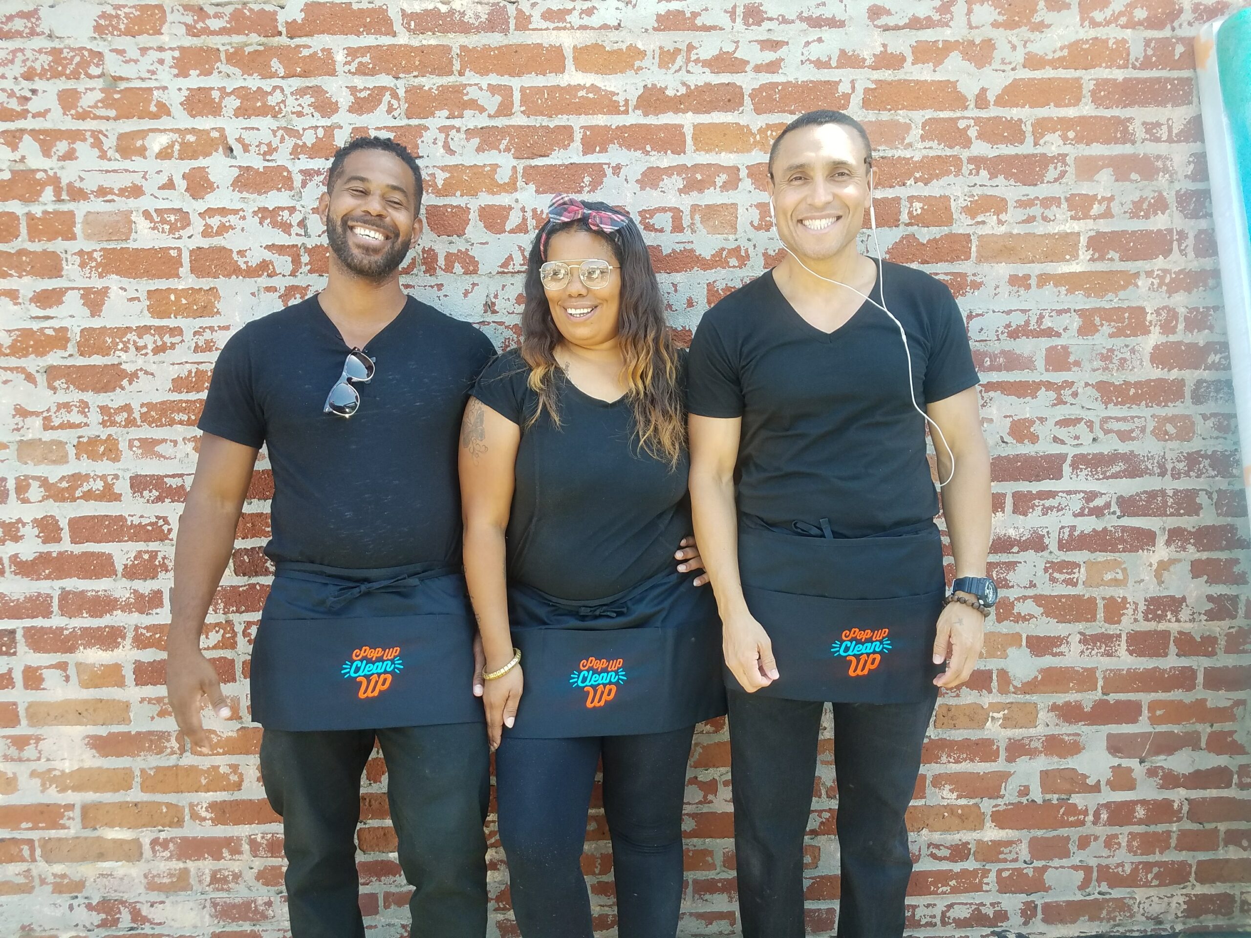 Three PopUP CleanUP Crew members in all black with branded aprons standing in front of a brick wall.