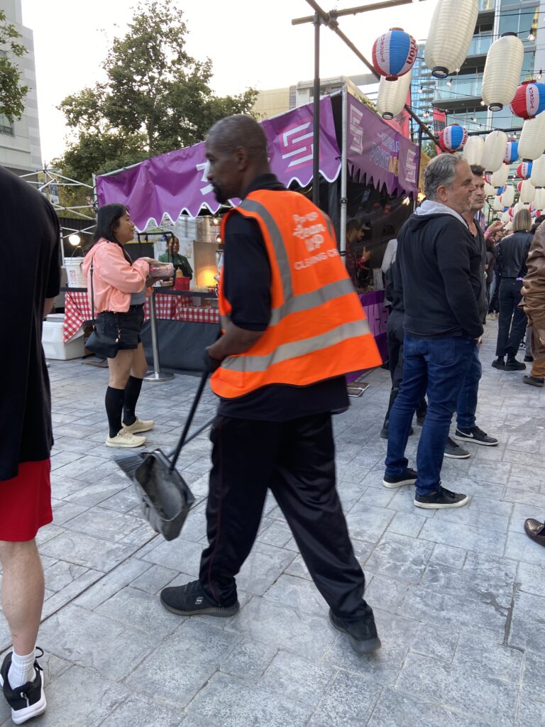 A PopUP CleanUP crew member with a broom and dustpan wearing an orange reflective branded vest outside at Trinity Auditorium Downtown Los Angeles for the Japanese Wrestling Event.