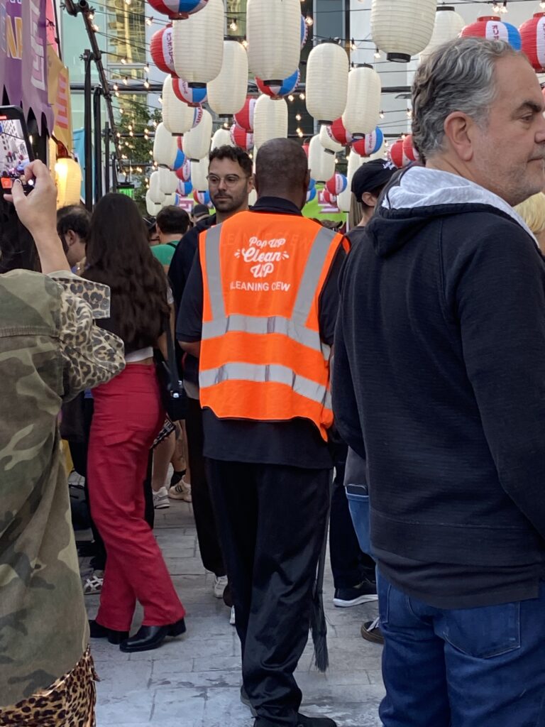 A live event porter with a neon orange PopUP CleanUP Cleaning Crew Vest working his way through the crowd at the Japanese Wrestling Event outside Trinity Auditorium in DTLA.