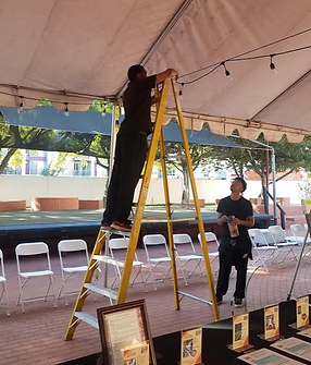 A popUP crew member on a ladder stringing lights under a tent in front of a row of folding white chairs.