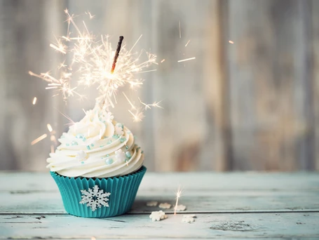 A white frosted cupcake with a blue wrapper decorated with a snowflake. A sparkler is lit up sticking out of the top of the frosting. The cupcake sits on a wooden farm table.