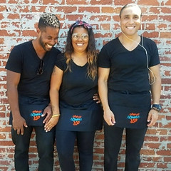 Three crew members smiling and laughing, standing against a brick wall with PopUP CleanUP aprons on