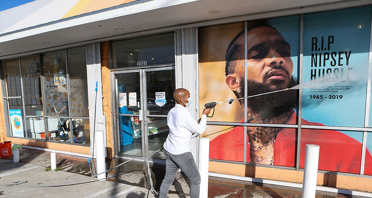 Our CEO Kenetia Lee powerwashing the windows of a local grocery store in the Crenshaw district.  Behind the window is a giant poster of Nipsey Hustle with the words RIP 1985-2019.