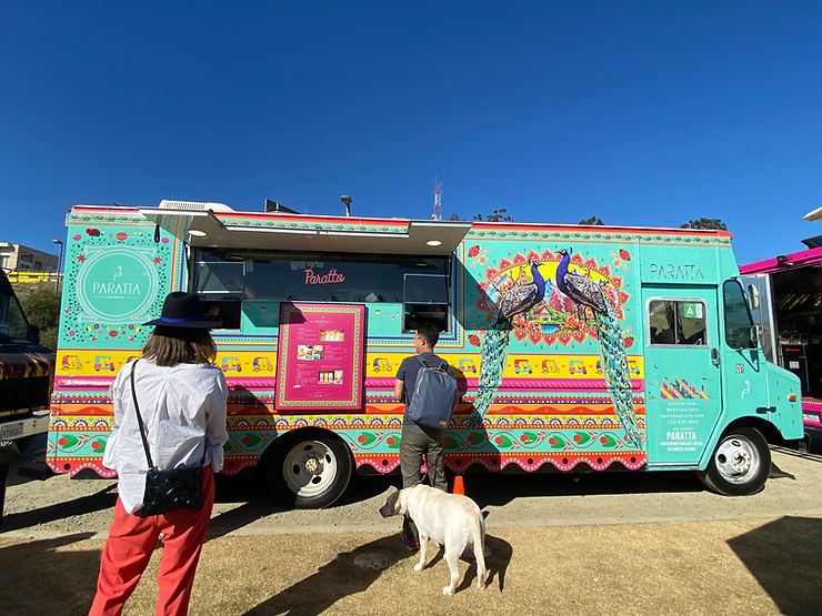 A turquoise and pink food truck with peacocks in the log at the Renegade Craft Fair.  Two patrons stand in line, one with a yellow Labrador.