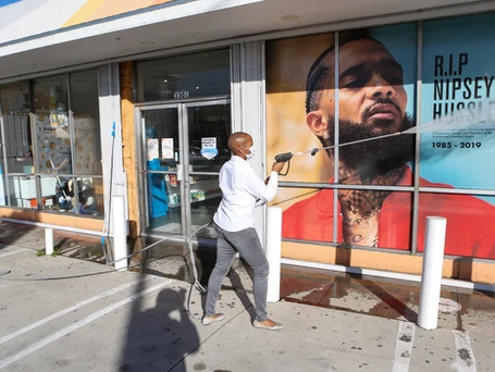 CEO Kenetia Lee power washing the outside windows of the Crenshaw District's Hank's Market. A poster with RIP Nipsey Hustle fills the windows being power washed.