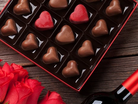 A box of chocolate hearts next to some red roses and a glimpse of a bottle of wine.