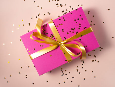 A pink gift box with a gold ribbon sprinkled with gold heart confetti.