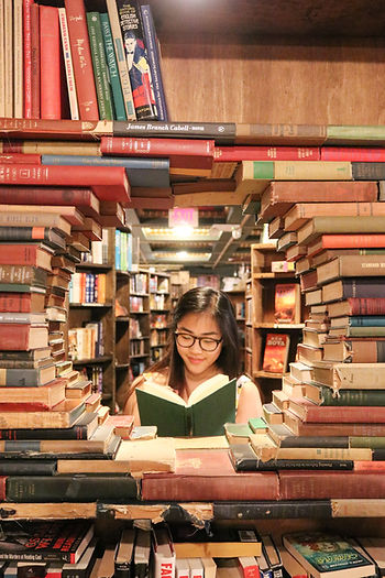 A woman with glasses seen reading through a portal of stacked books in a library. 