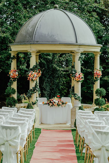 A wedding gazebo decorated with vines and bouquets of peach and white roses in front of a pink aisle. Wood chairs wrapped in white cloth line either side of the aisle.