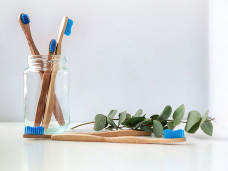 Three bamboo tooth brushes with blue bristles in a glass jar. Two bamboo tooth brushes lying on a table next to a eucalyptus branch.