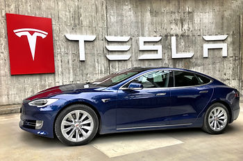 A blue Tesla electric car in front of the Tesla logo and sign.