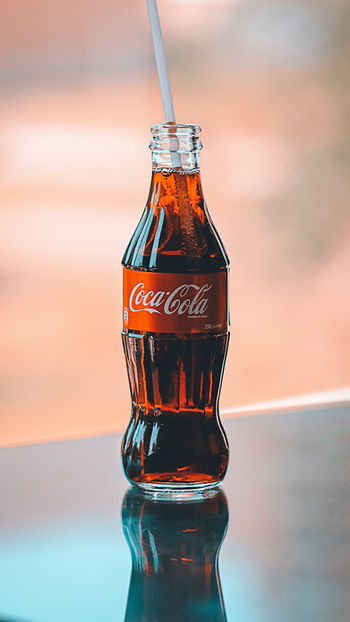 A glass bottle of Coca-Cola with a straw in it on a black reflective surface.