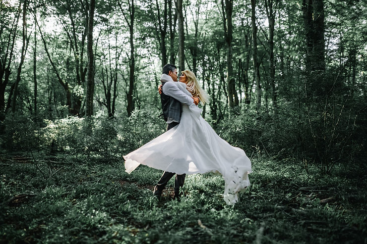 A happy couple embracing.  The man is lifting the woman into a twirl so that her white wedding dress sprays around.  They are standing in the middle of green woods.