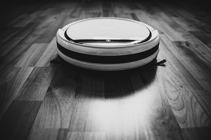 A robo-vacuum disk sliding along a wood floor in a black and white photograph.