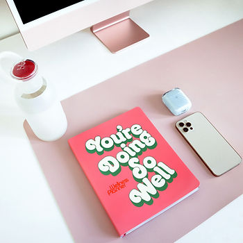 A book on a desk with the title YOU'RE DOING SO WELL. next to an iPhone, a water bottle, and a desktop computer.