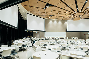 A circular corporate meeting room with round white tables and white and black chairs.  Hanging down from the ceiling are multiple screens for projectors and the ceiling has wooden tiles that pull down with hidden projectors aimed at the screens.