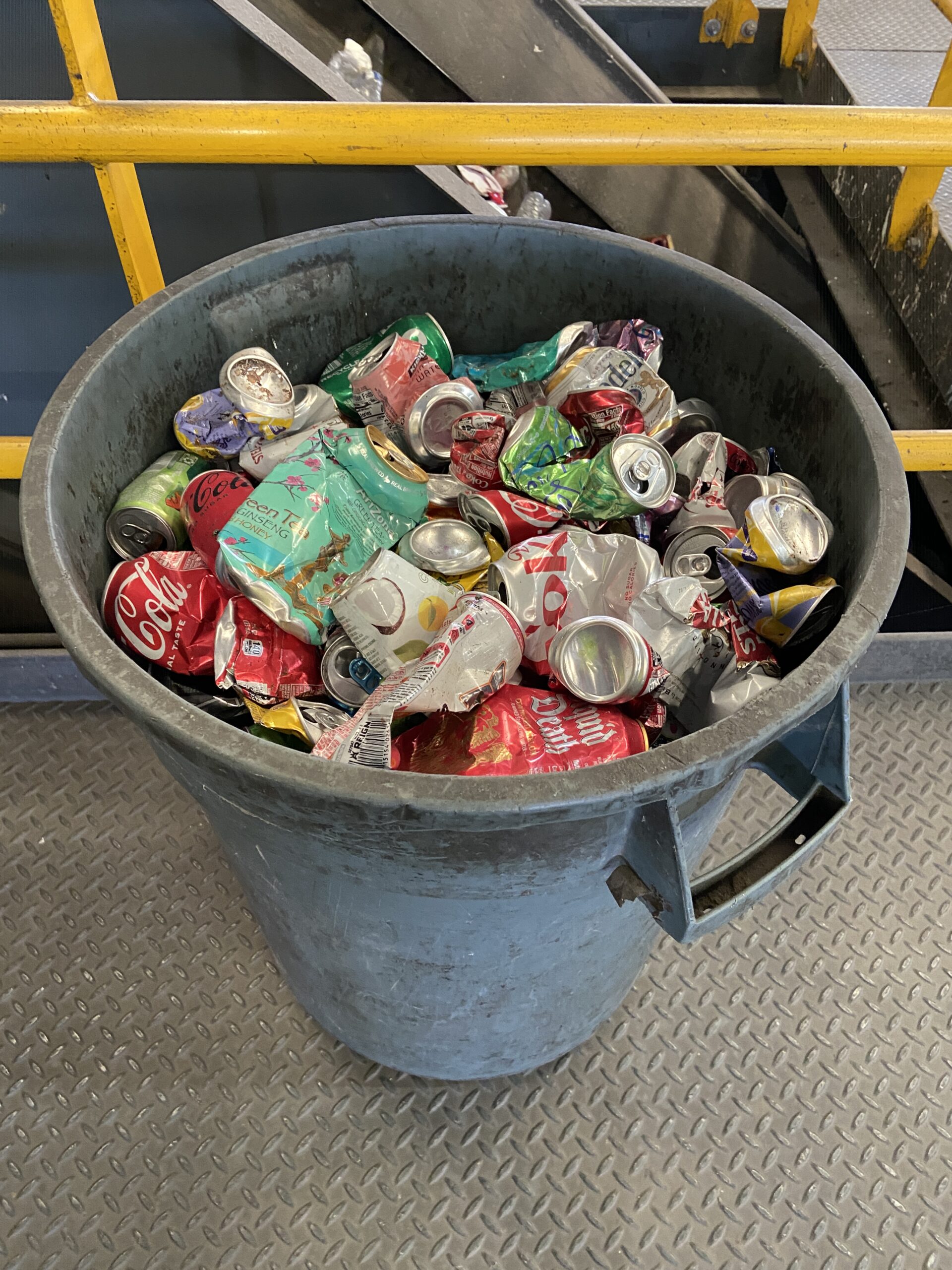 A grey plastic 50 gallon trash can filled with aluminum cans.