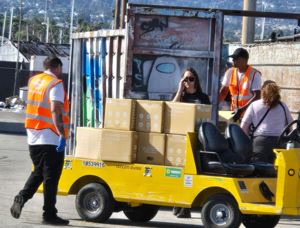 Two PopUP CleanUP crew members on the utility cart moving boxes of toss ware around the lot in front of a graffiti-tagged dumpster at the Port of Los Angeles in San Pedro.